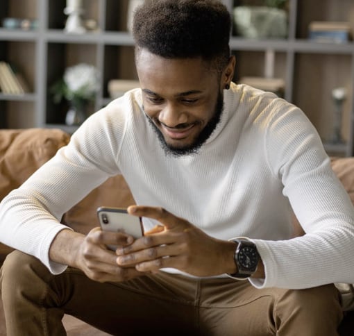 A man with a beard and short hair is sitting on a couch, smiling while looking at his smartphone. He is wearing a white sweater, brown pants, and a black smartwatch. The background shows shelves with books and decorative items.