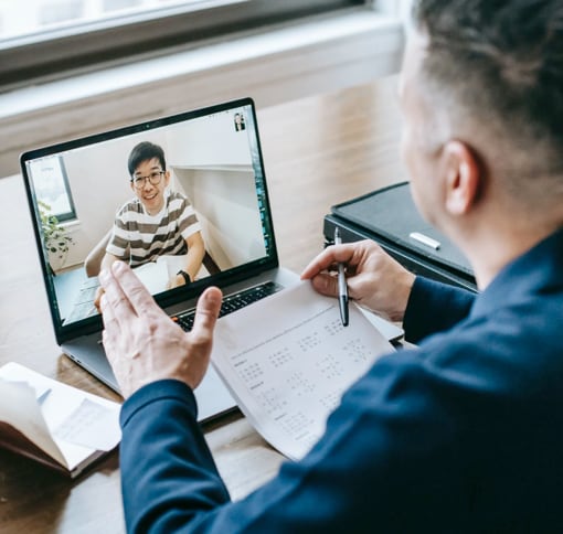 A man in a blue shirt is on a video call using a laptop. He holds a document with graphs while talking to another man, visible on the laptop screen, wearing glasses and a striped shirt. The setting is a desk with a briefcase and papers around.