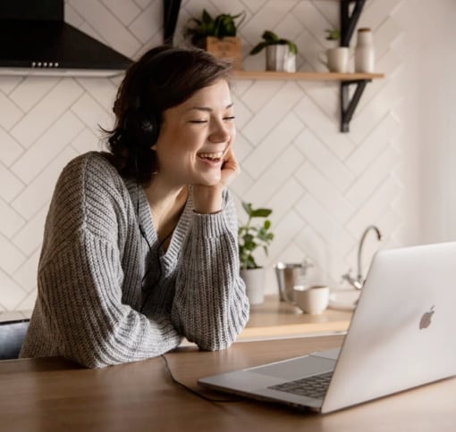 A woman with short dark hair and a gray sweater is smiling and leaning her head on her hand while on a video call. She sits at a kitchen table with an open laptop in front of her. The background shows a modern kitchen with white tiled walls and shelves.