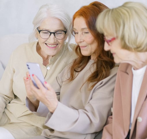 Three elderly women are sitting closely together, smiling and looking at a smartphone held by the woman in the middle. One has white hair and glasses, another has red hair, and the third has blonde hair and wears a light brown jacket. They appear to be enjoying a moment together.