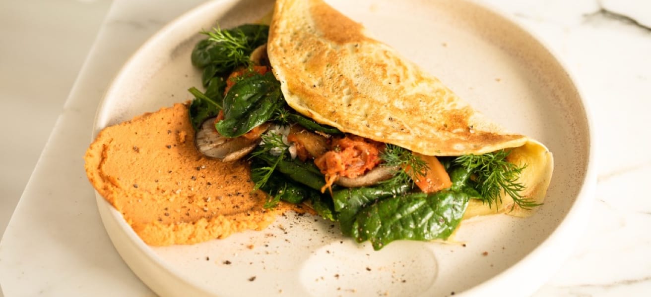 A folded omelette on a beige plate, filled with fresh spinach, grilled vegetables, and herbs. Beside the omelette is a dollop of a reddish spread, likely a hummus or puree, sprinkled with black pepper